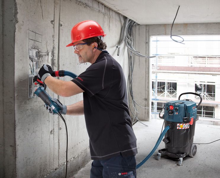 บุคคลที่สวมอุปกรณ์ความปลอดภัยกำลังบดผนังคอนกรีต โดยมีเครื่องดูดฝุ่นทำงานอยู่บริเวณใกล้เคียง