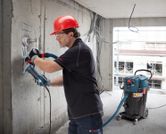 บุคคลที่สวมอุปกรณ์ความปลอดภัยกำลังบดผนังคอนกรีต โดยมีเครื่องดูดฝุ่นทำงานอยู่บริเวณใกล้เคียง