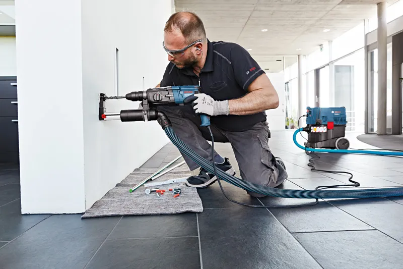 บุคคลที่สวมอุปกรณ์ด้านความปลอดภัยกำลังเจาะเข้าไปในผนังที่มีอุปกรณ์ดูดฝุ่น.