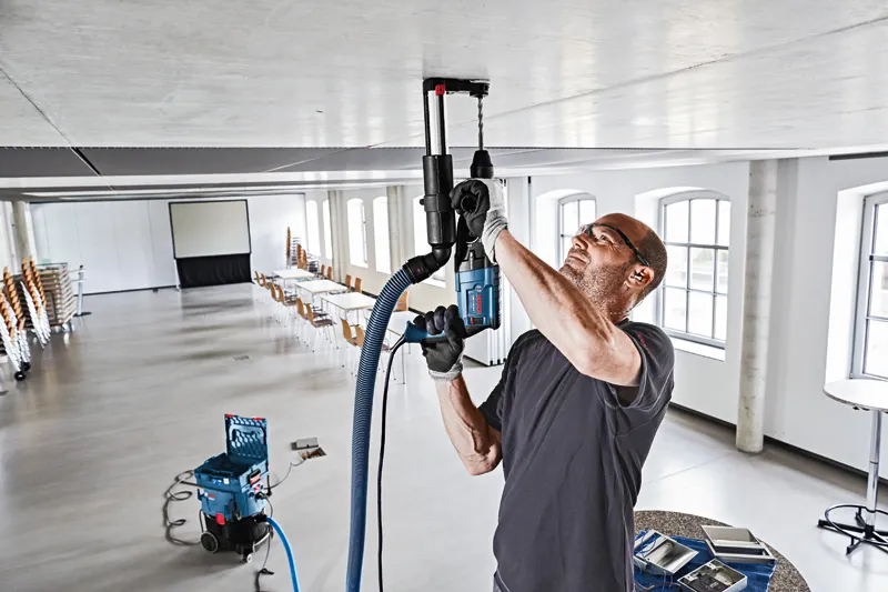 บุคคลที่สวมอุปกรณ์ด้านความปลอดภัยกำลังเจาะเพดานโดยใช้ระบบดูดฝุ่น.