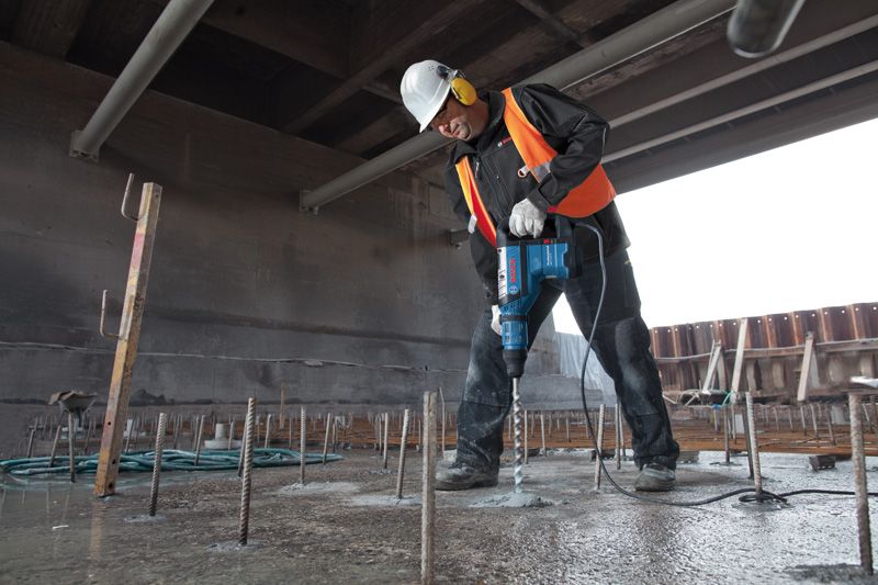 บุคคลที่สวมอุปกรณ์ความปลอดภัยกำลังเจาะเข้าไปในคอนกรีตด้วยค้อนโรตารี่ที่ไซต์ก่อสร้าง.