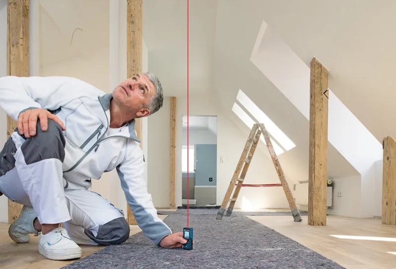 บุคคลใช้เครื่องวัดเลเซอร์เพื่อตรวจสอบการจัดแนวแนวตั้งในห้องใต้หลังคาที่มีแสงสว่าง