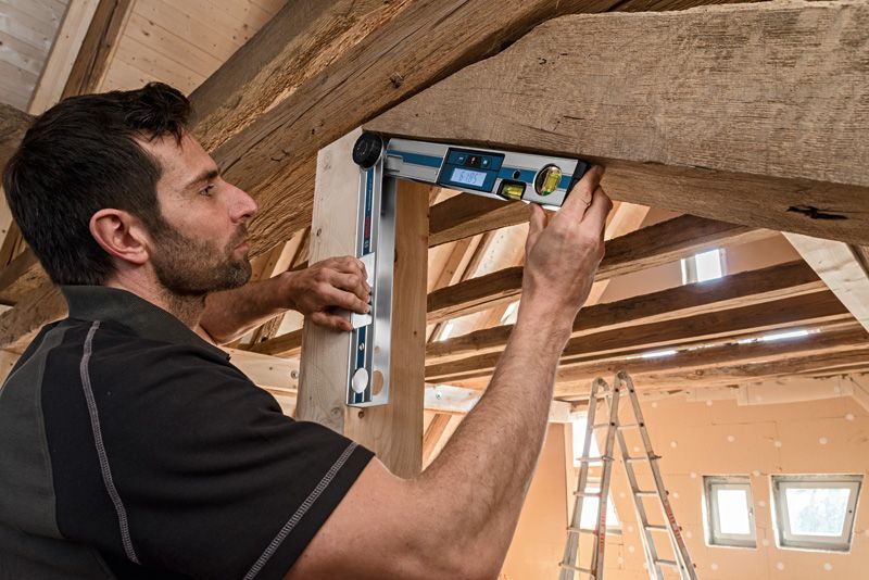 ผู้คนกำลังตรวจสอบมุมลำแสงไม้โดยใช้เครื่องวัดความเอียงแบบดิจิตอลใต้หลังคาไม้.