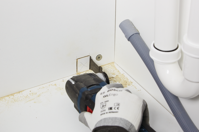 บุคคลที่สวมอุปกรณ์ความปลอดภัยกำลังตัดช่องสี่เหลี่ยมในผนังตู้ใกล้กับท่อ