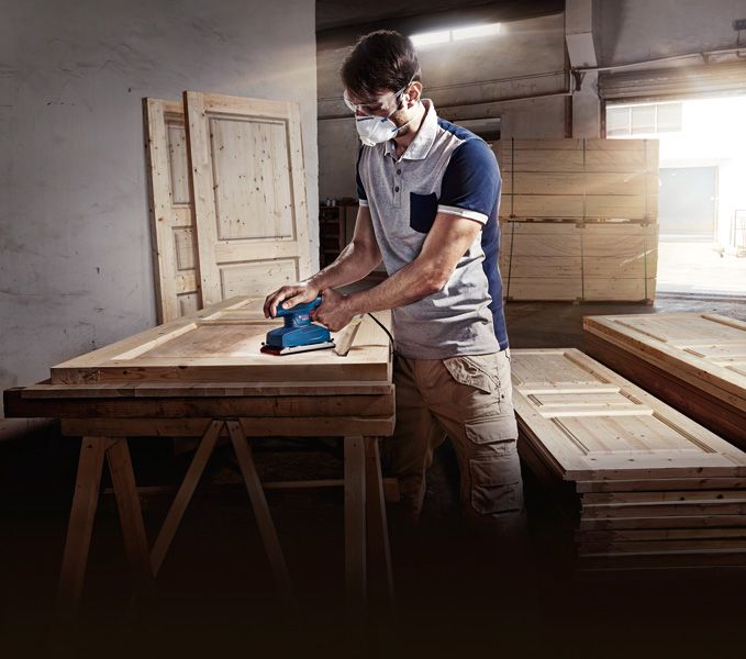 บุคคลที่สวมอุปกรณ์ความปลอดภัยกำลังขัดประตูไม้ด้วยเครื่องขัดแบบวงโคจร