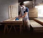 บุคคลที่สวมอุปกรณ์ความปลอดภัยกำลังขัดประตูไม้ด้วยเครื่องขัดแบบวงโคจร