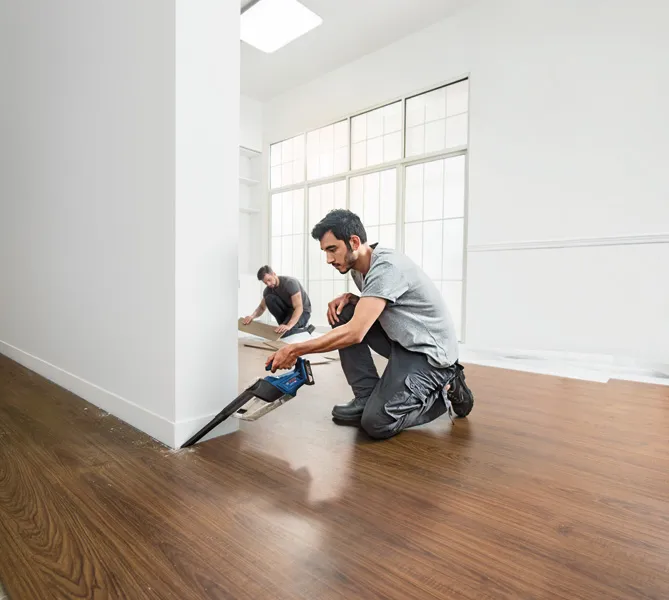 คนๆ หนึ่งกำลังตัดพื้นบริเวณใกล้กำแพงด้วยเลื่อยไร้สาย ขณะที่อีกคนหนึ่งกำลังติดตั้งแผงพื้น.