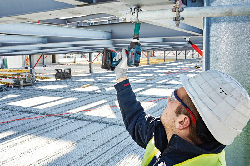 บุคคลที่สวมอุปกรณ์ด้านความปลอดภัยใช้ประแจกระแทกไร้สายกับคานโลหะ.