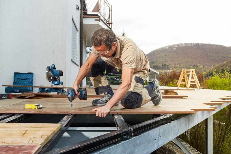 บุคคลที่สวมอุปกรณ์ด้านความปลอดภัยใช้ไขควงกระแทกไร้สายในการยึดพื้นไม้ระเบียง.