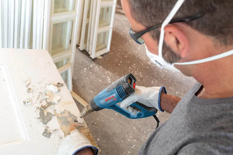 บุคคลที่สวมอุปกรณ์ด้านความปลอดภัยใช้ปืนเป่าลมร้อนเพื่อลอกสีออกจากประตู