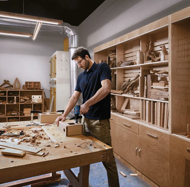 ผู้ที่สวมอุปกรณ์ด้านความปลอดภัยใช้สว่านไร้สายบนกล่องไม้ในโรงงาน.