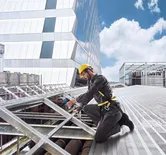 ผู้ที่สวมอุปกรณ์ด้านความปลอดภัยใช้สว่านไร้สายบนโครงหลังคาโลหะ.