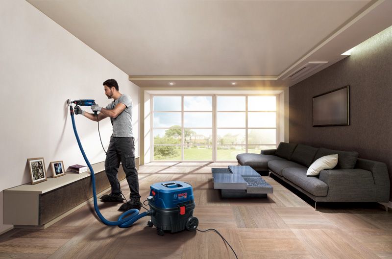 คนๆ หนึ่งกำลังเจาะผนังห้องนั่งเล่นโดยใช้เครื่องมือไฟฟ้าที่ต่อกับเครื่องดูดฝุ่น.