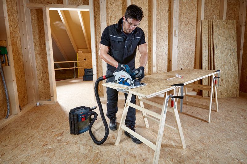 บุคคลที่สวมอุปกรณ์ด้านความปลอดภัยกำลังตัดไม้อัดด้วยเลื่อยวงเดือนที่เชื่อมต่อกับเครื่องดูดฝุ่น.