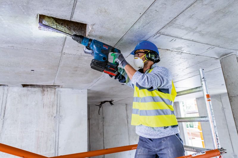 บุคคลที่สวมอุปกรณ์ด้านความปลอดภัยกำลังเจาะเข้าไปในเพดานคอนกรีตด้วยสว่านโรตารี่ไร้สาย.