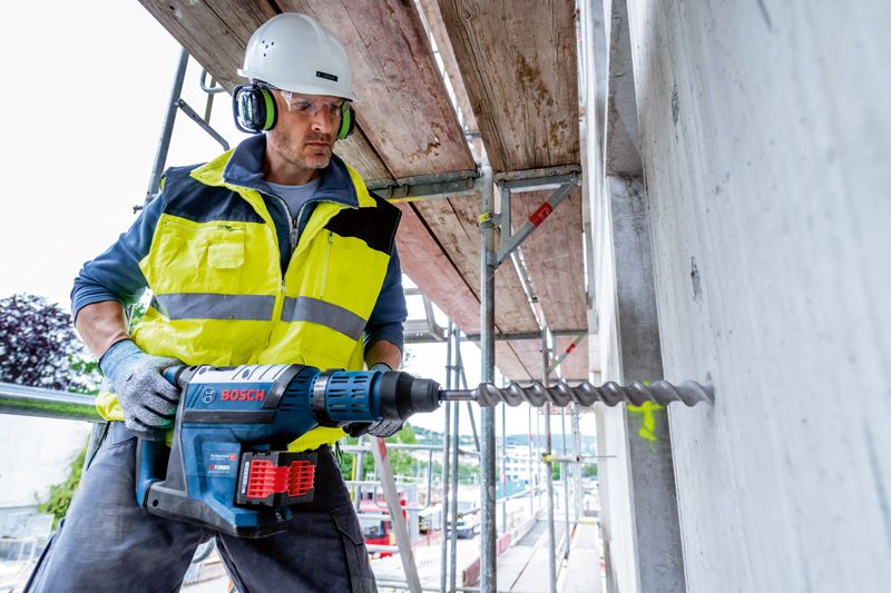 บุคคลที่สวมอุปกรณ์ด้านความปลอดภัยกำลังเจาะเข้าไปในคอนกรีตด้วยสว่านโรตารี่ไร้สาย.