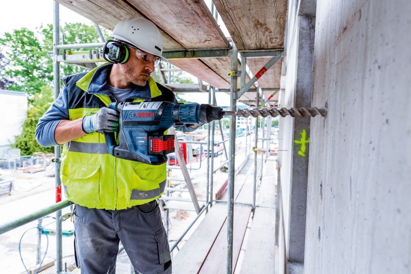 บุคคลที่สวมอุปกรณ์ความปลอดภัยกำลังเจาะเข้าไปในผนังคอนกรีตในพื้นที่ก่อสร้าง.