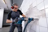 ผู้ที่สวมอุปกรณ์ด้านความปลอดภัยกำลังถอดกระเบื้องผนังโดยใช้สว่านโรตารี่ไร้สาย.