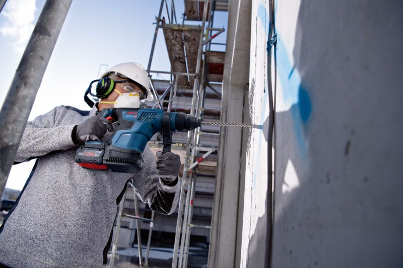 บุคคลที่สวมอุปกรณ์ความปลอดภัยกำลังเจาะเข้าไปในผนังคอนกรีตในพื้นที่ก่อสร้าง.