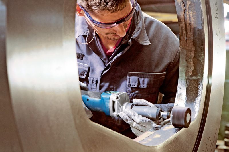 บุคคลที่สวมอุปกรณ์ด้านความปลอดภัยกำลังขัดวงแหวนโลหะขนาดใหญ่ด้วยเครื่องเจียรตรงแบบไร้สาย.