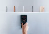 บุคคลหนึ่งสแกนผนัง drywall ด้วยเครื่องตรวจจับเพื่อระบุท่อและสายไฟที่ซ่อนอยู่.