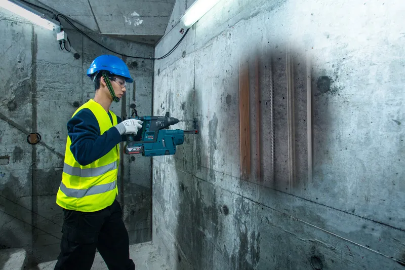 บุคคลที่สวมอุปกรณ์ด้านความปลอดภัยสแกนผนังคอนกรีตด้วยเครื่องตรวจจับ ซึ่งเผยให้เห็นท่อที่อยู่ด้านใน.