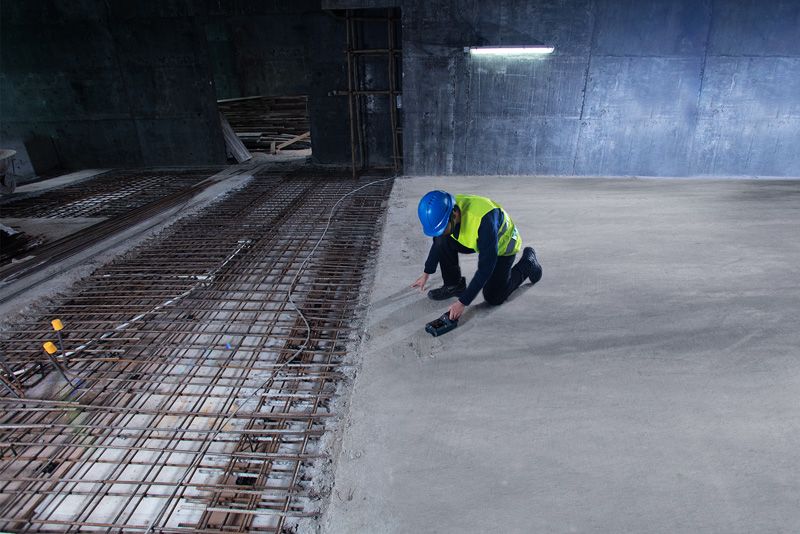 บุคคลที่สวมอุปกรณ์ด้านความปลอดภัยกำลังสแกนคอนกรีตด้วยเครื่องตรวจจับบริเวณใกล้เหล็กเส้นที่โผล่ออกมา.