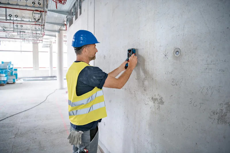 บุคคลที่สวมอุปกรณ์ด้านความปลอดภัยสแกนผนังคอนกรีตด้วยอุปกรณ์ตรวจจับ.