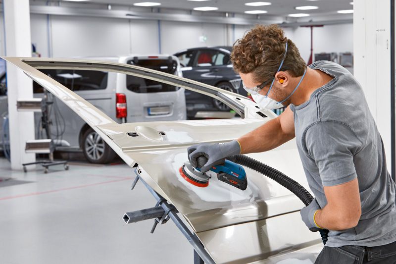 บุคคลที่สวมอุปกรณ์ความปลอดภัยกำลังขัดประตูรถด้วยเครื่องขัดไฟฟ้าในอู่ซ่อมรถยนต์.