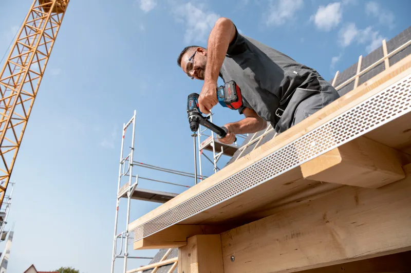 บุคคลที่สวมอุปกรณ์ความปลอดภัยกำลังเจาะเข้าไปในคานไม้ของอาคารที่กำลังก่อสร้าง.