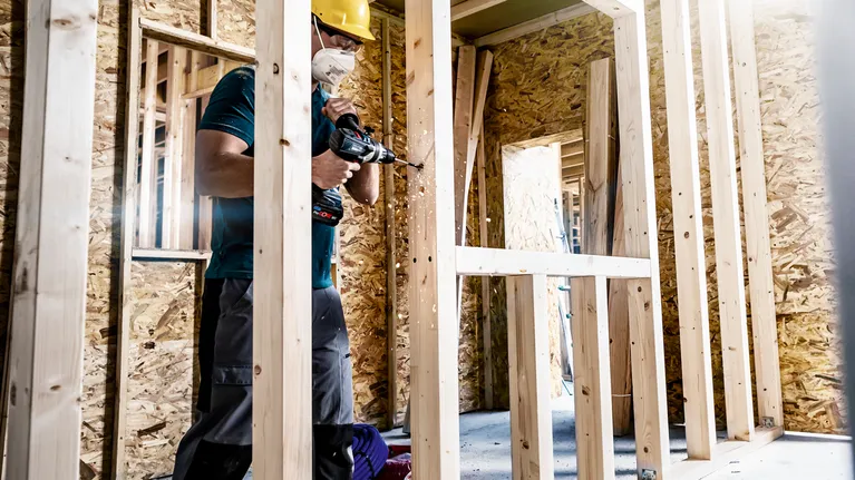 คนสวมอุปกรณ์ความปลอดภัยกำลังเจาะเข้าไปในโครงไม้ที่ไซต์ก่อสร้าง