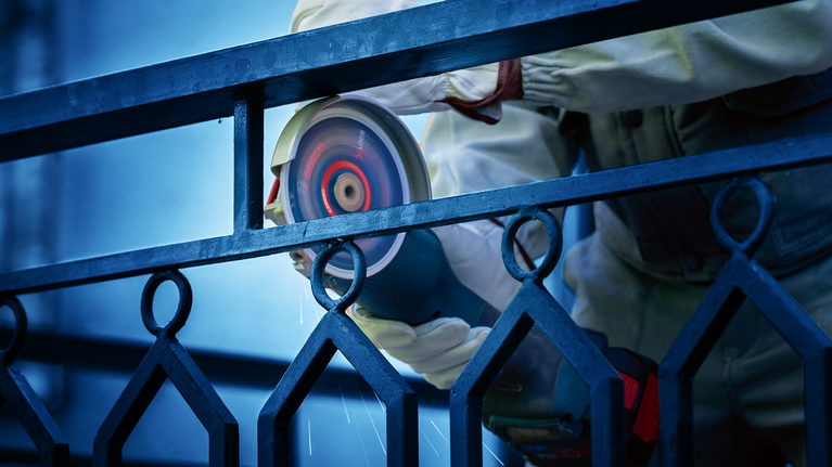 บุคคลที่สวมอุปกรณ์ความปลอดภัยกำลังเจียรรั้วโลหะด้วยเครื่องมือไฟฟ้า