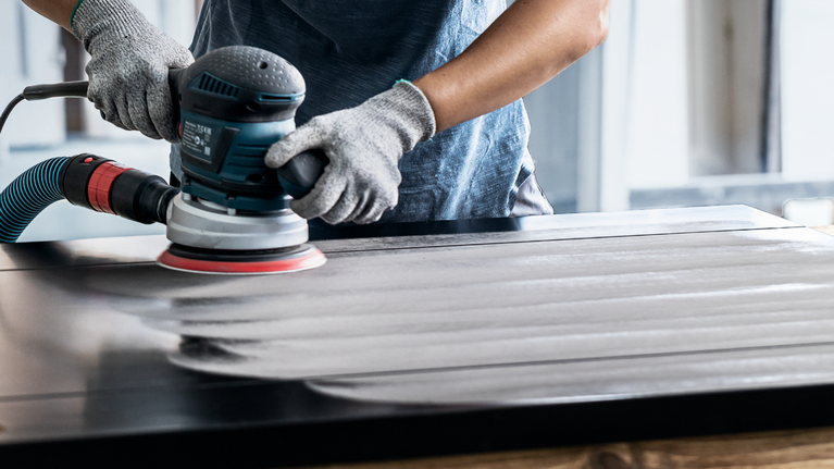 บุคคลที่สวมอุปกรณ์ด้านความปลอดภัยกำลังขัดพื้นผิวไม้ด้วยเครื่องมือไฟฟ้า