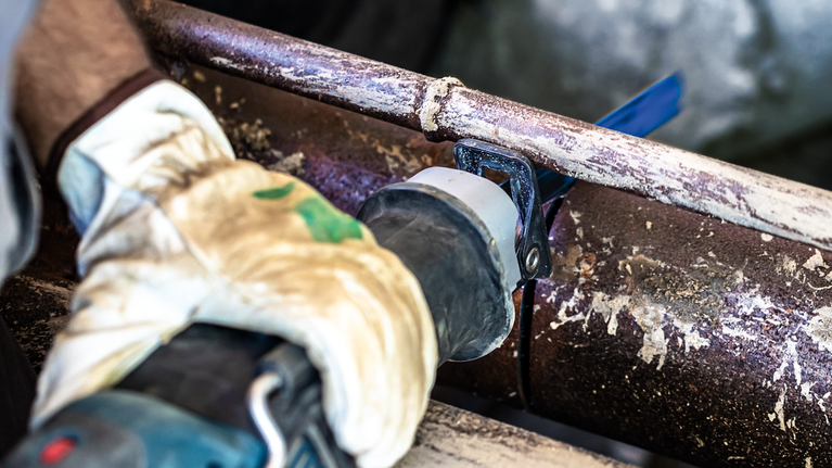 ผู้ที่สวมอุปกรณ์ป้องกันความปลอดภัยกำลังตัดท่อโลหะที่เป็นสนิมด้วยเลื่อยตัดโลหะแบบลูกสูบ