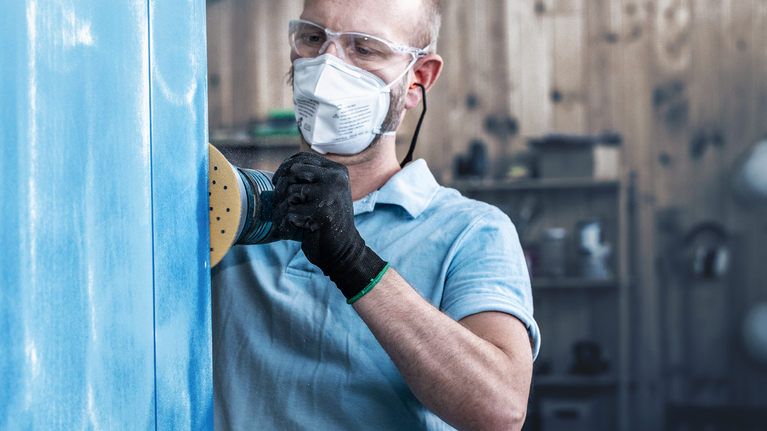 บุคคลที่สวมอุปกรณ์ด้านความปลอดภัยกำลังขัดแผงสีน้ำเงินด้วยเครื่องขัดไฟฟ้า