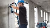 บุคคลที่สวมอุปกรณ์ความปลอดภัยกำลังขัดผนังคอนกรีตโดยมีเครื่องดูดฝุ่นอยู่ใกล้ๆ