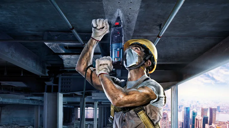 บุคคลที่สวมอุปกรณ์ความปลอดภัยกำลังเจาะเข้าไปในเพดานคอนกรีตในพื้นที่ก่อสร้าง.