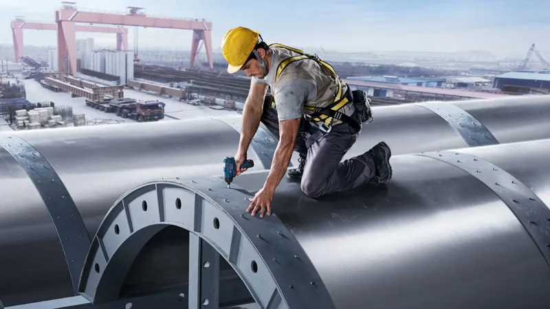 บุคคลที่สวมอุปกรณ์ด้านความปลอดภัยกำลังขันน็อตบนโครงสร้างโลหะขนาดใหญ่โดยใช้เครื่องมือไฟฟ้า.