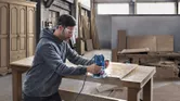 ผู้ที่สวมอุปกรณ์ด้านความปลอดภัยกำลังใช้เราเตอร์บนโต๊ะไม้ในโรงงาน.