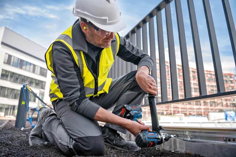 บุคคลที่สวมอุปกรณ์ด้านความปลอดภัยใช้สว่านกระแทกไร้สายบนโครงรั้วโลหะ.