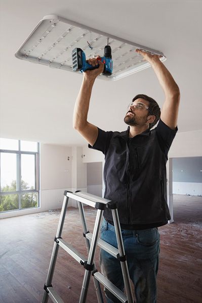 บุคคลที่สวมอุปกรณ์ด้านความปลอดภัยติดตั้งโคมไฟเพดานโดยใช้สว่านไฟฟ้า.