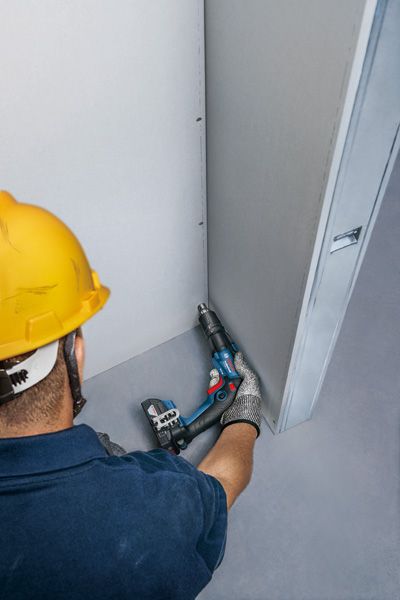 บุคคลที่สวมอุปกรณ์ด้านความปลอดภัยกำลังยึดผนังยิปซั่มด้วยไขควงไร้สาย.