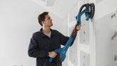 คนๆ หนึ่งกำลังขัดแผ่นผนัง drywall ด้วยเครื่องขัด drywall แบบไฟฟ้ากับผนังที่ยังไม่เสร็จ