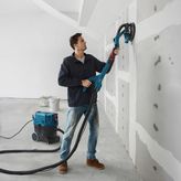 คนๆ หนึ่งกำลังขัดผนัง drywall โดยใช้เครื่องขัด drywall ที่ต่อกับเครื่องดูดฝุ่น