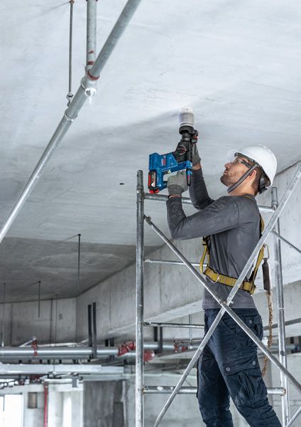 บุคคลที่สวมอุปกรณ์ความปลอดภัยยืนอยู่บนนั่งร้านโดยใช้สว่านไฟฟ้าบนเพดาน.