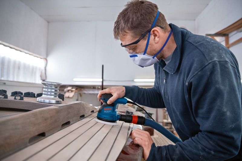 บุคคลที่สวมอุปกรณ์ป้องกันความปลอดภัยกำลังขัดแผ่นไม้ด้วยเครื่องขัดแบบสุ่มวงโคจร.