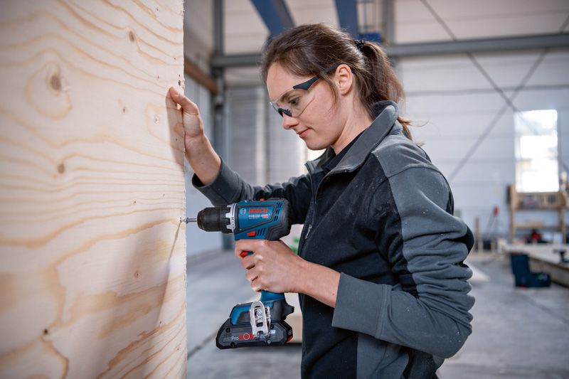 บุคคลที่สวมอุปกรณ์ความปลอดภัยกำลังเจาะลงบนแผ่นไม้ในโรงงาน.