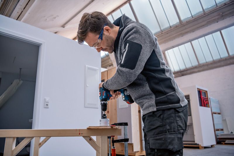 บุคคลที่สวมอุปกรณ์ด้านความปลอดภัยกำลังเจาะรูในไม้โดยใช้สว่านไฟฟ้าไร้สาย.