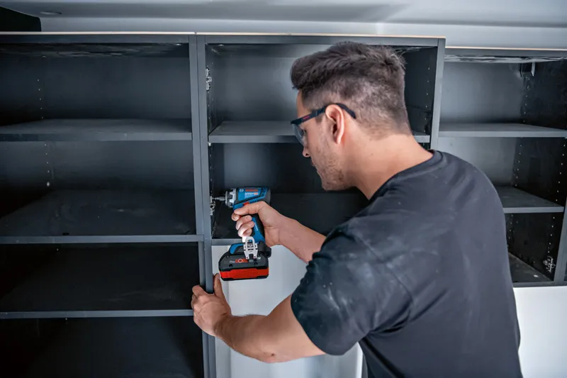 บุคคลที่สวมอุปกรณ์ด้านความปลอดภัยกำลังประกอบชั้นวางด้วยสว่านหรือไขควงไร้สาย.