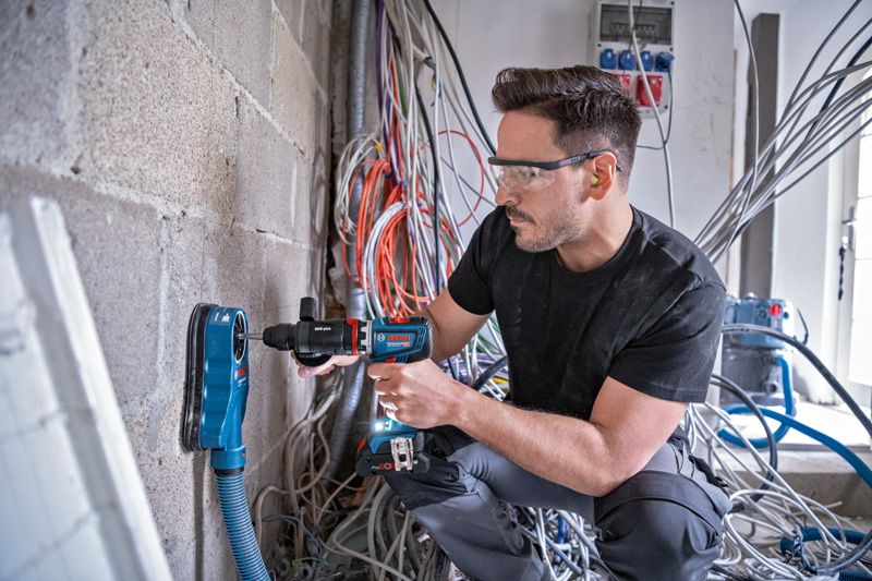 บุคคลที่สวมอุปกรณ์ความปลอดภัยกำลังเจาะเข้าไปในผนังคอนกรีตโดยใช้เครื่องดูดฝุ่น.